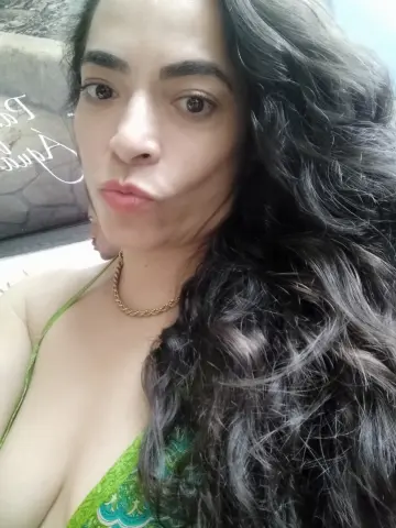 SweetsAnne pouts her lips while showing off her long, wavy dark hair and gold chain necklace, wearing a green patterned bikini top.