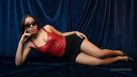 Stylish woman in a red leather bodysuit and black skirt posing against a dark blue velvet backdrop.