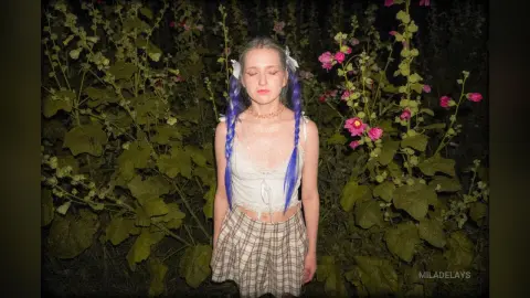 Dreamy woman with blue braids in a white tank top standing amidst lush green garden flowers at night.