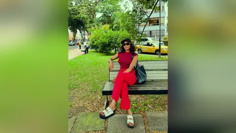 Stylish woman in red jumpsuit and black hat sitting on a park bench on a sunny day.