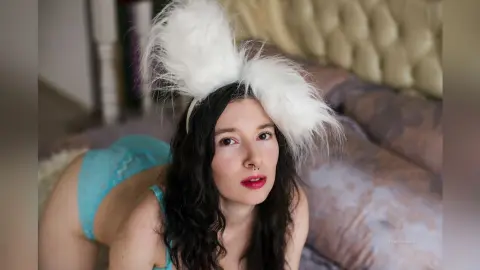 Young woman with dark hair wearing white fuzzy bunny ears and blue lace lingerie on a bed.