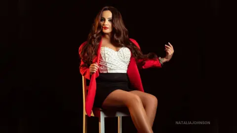 Elegant woman in a white corset and red blazer sitting on a chair against a black studio background.