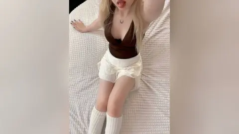 LillyEyes licks her lips while wearing a brown bodysuit and white skirt, lying back on a white textured bed.