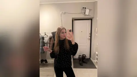 Young woman in a black polka dot sweater taking a mirror selfie in a minimalist modern apartment.