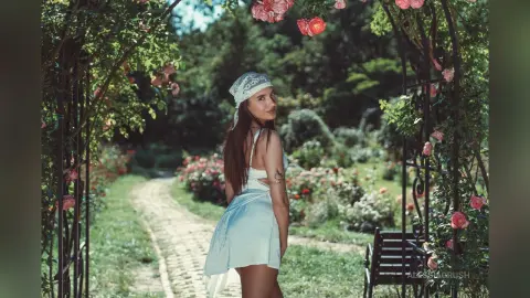 AlessiaCrush poses in a light blue sundress and white lace headscarf on a garden path surrounded by roses.