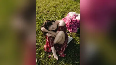 Dreamy portrait of a woman in a red floral dress sitting on green grass with pink flowers.