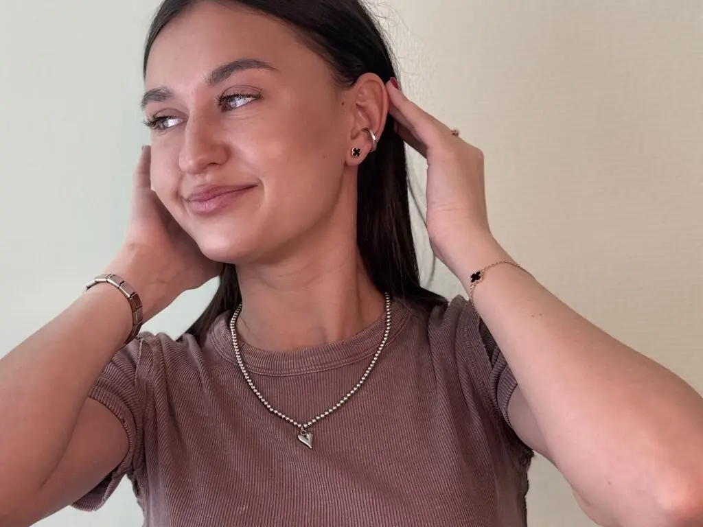 Smiling young woman in a brown ribbed t-shirt wearing silver jewelry against a neutral background.