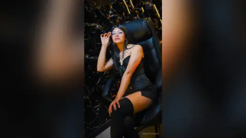 Stylish woman in a black lace outfit and glasses posing confidently in a dark, moody luxury setting.