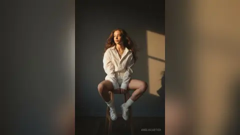 Young woman in white button-down shirt and shorts sitting in moody sunlight against a gray wall.