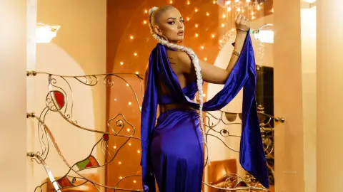 Anastasia poses in a blue satin outfit with long hanging sleeves, showing her blonde braided hair and bare back.