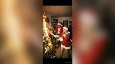 Woman in a red velvet Santa outfit celebrating with a person in a Shrek costume by a glowing Christmas tree.