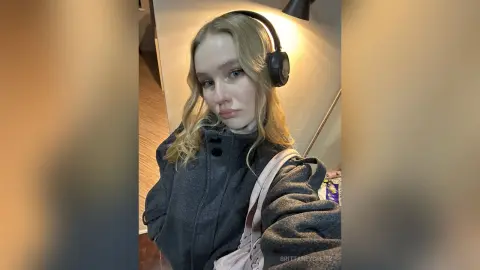 Blonde woman in a cozy grey wool coat and black headphones relaxing in a warm indoor setting.