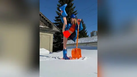 Cheerful woman in blue winter gear holding a boot and orange snow shovel in a sunny snowy yard.