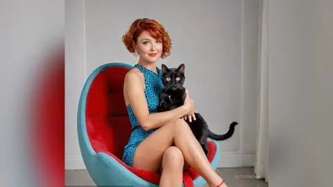 Redheaded woman in a blue polka dot dress smiling while holding a black cat in a modern red lounge chair.