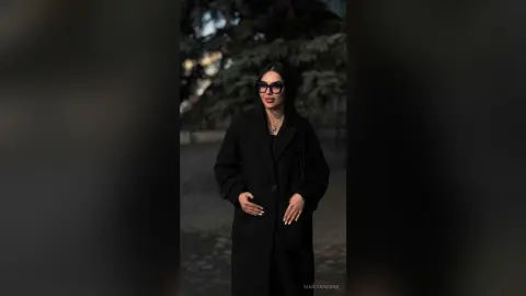 Elegant woman in a black winter coat and glasses standing in a moody, dark park setting.