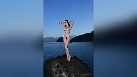 Slim woman in a silver bikini posing on dark rocks by the ocean under a clear blue twilight sky.