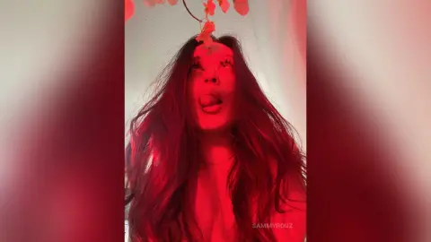 SammyRouz poses with her long dark hair down, sticking her tongue out under a red light.