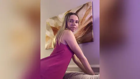 Young woman in a magenta sleeveless dress posing in a cozy room with warm lighting and abstract wall art.