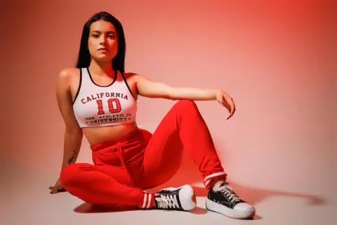 Young woman in white California University crop top and red sweatpants sitting against a warm studio background.