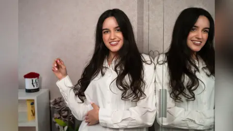 Smiling young woman with long dark hair wearing a white button-down shirt in a bright indoor setting.