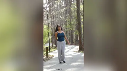 Smiling young woman in a blue tank top and grey sweatpants walking on a sunny forest path.