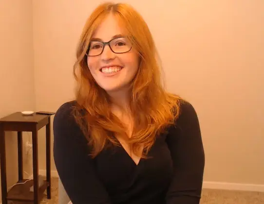 GoodluckAnne smiles with her long red hair down, wearing a black long-sleeved top in a simple indoor setting.