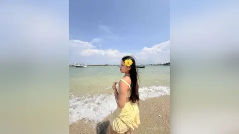 Young woman in a yellow sundress with a yellow flower in her hair standing on a sunny beach by the ocean waves.