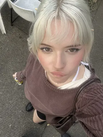 Blonde woman in a brown sweater and white tank top sitting outdoors on a sunny day.