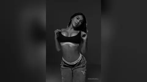 Glamorous woman in black Calvin Klein bralette and denim jeans posing in a moody black and white studio portrait.