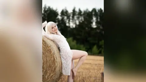 Blonde woman in a white lace dress leaning against a hay bale in a dreamy rural field setting.