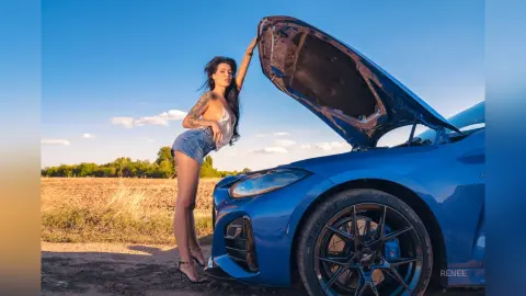 Fashionable woman in denim shorts posing by a blue sports car with an open hood in a sunny field.