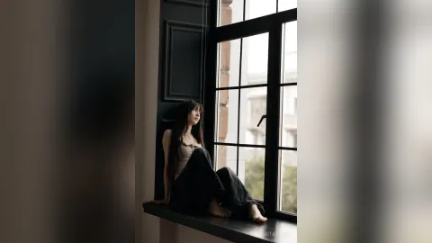 MintaTukes — MintaTukes sits on a window sill in a black tank top and black pants, looking out the window.