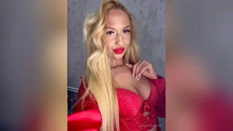 Blonde woman in a glamorous red satin outfit posing with a confident expression against a grey background.