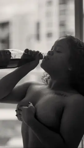 Nude woman drinking from a glass bottle by a window in a moody, black and white cinematic setting.