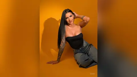 Smiling woman in a black tube top and grey jeans posing against a vibrant yellow studio background.