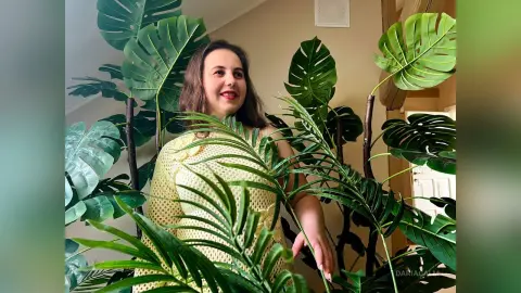 DariaCalm smiles while standing behind large green monstera and palm leaves in an indoor setting, wearing a light yellow mesh top.