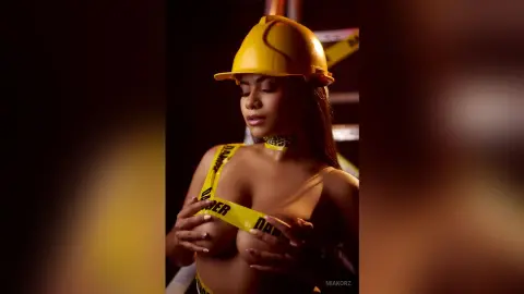 MiaKorz wears a yellow hard hat and yellow caution tape across her bare breasts in a dark setting.