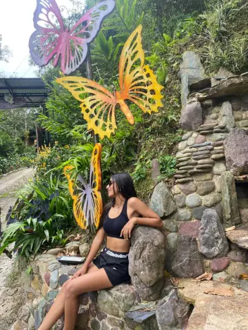 CeleGarcia sits on a stone wall outdoors, wearing a black crop top and black shorts, surrounded by large colorful butterfly decorations.