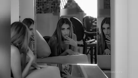 Melancholic black and white mirror selfie of a young woman in a casual top looking thoughtful in a bright room.