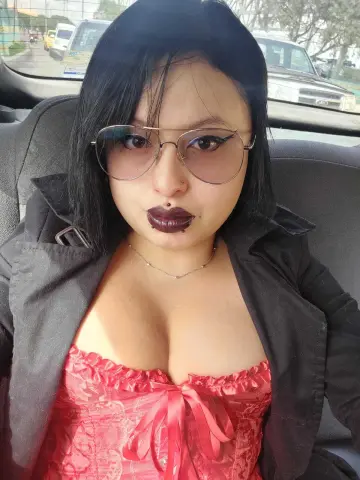 Noirdesire660 wears dark lipstick, glasses, and a red corset under a black jacket while sitting in a car.