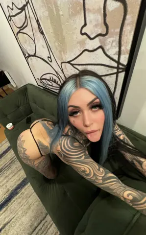 Tattooed woman with blue hair sitting on a green velvet sofa in front of large abstract line art.