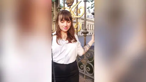 StacyMalvin poses outdoors wearing a white button-down shirt and a black skirt, standing next to a decorative gold and black metal gate.