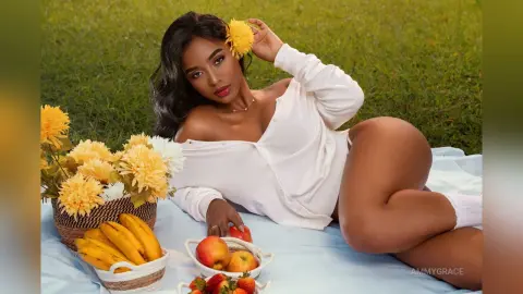 Beautiful woman in white blouse relaxing on a picnic blanket with flowers and fruit in a sunny meadow.