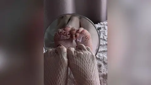 Close-up of feet in white fishnet tights reflected in a round mirror on a cozy textured rug.