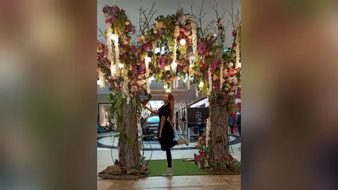 TiffanyAddams stands next to a large floral archway decorated with pink and white flowers, wearing a black dress and white sneakers.