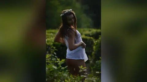 Young woman in a white lace camisole and flower crown posing in a lush green garden during golden hour.