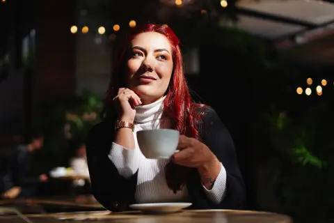 Red-haired woman in a white turtleneck enjoying coffee at a cozy, dimly lit outdoor cafe at night.