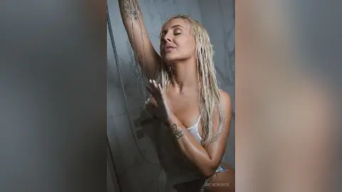 Anita poses wet in a shower with her blonde hair damp and her eyes closed, showing off her tattoos and bare skin.