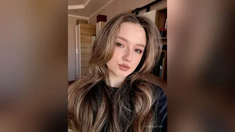 Young woman with long wavy brown hair wearing a black top in a cozy indoor setting.