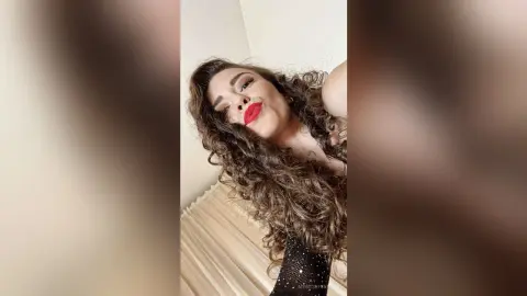 MontseFerrera smiles with red lipstick while wearing a black mesh top and letting her long, curly brown hair fall over her shoulders.